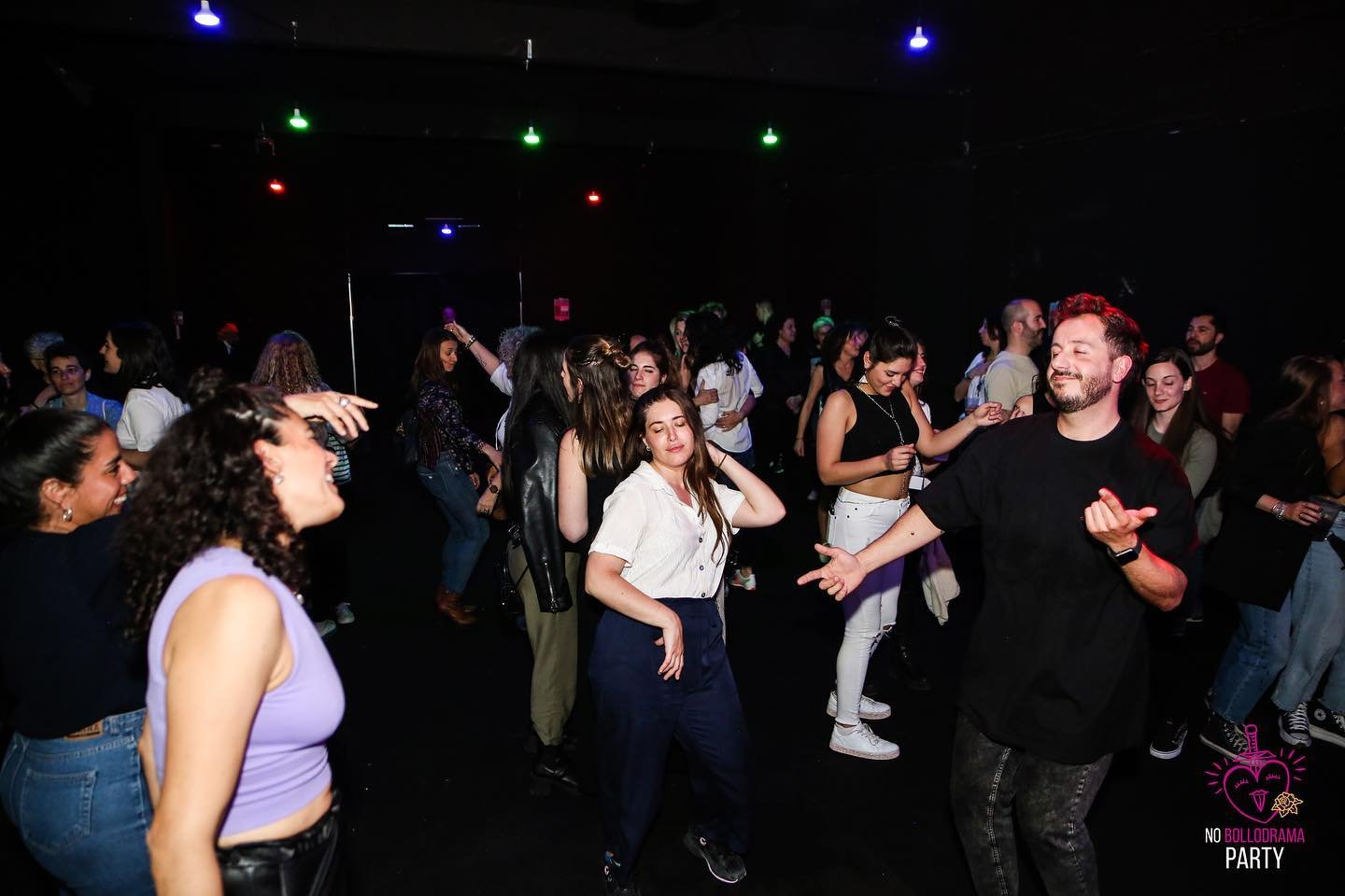 Varias personas bailan en un espacio oscuro con luces de colores suspendidas en el techo. Algunos parecen conversar o disfrutar la música mientras se mueven por la pista de baile.