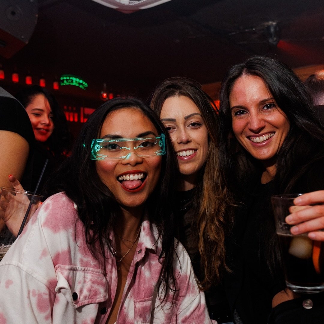 Tres mujeres sonríen a la cámara en un ambiente de discoteca con luces tenues rojas. La mujer a la izquierda lleva gafas de neon verde y una chaqueta rosa, sacando la lengua.