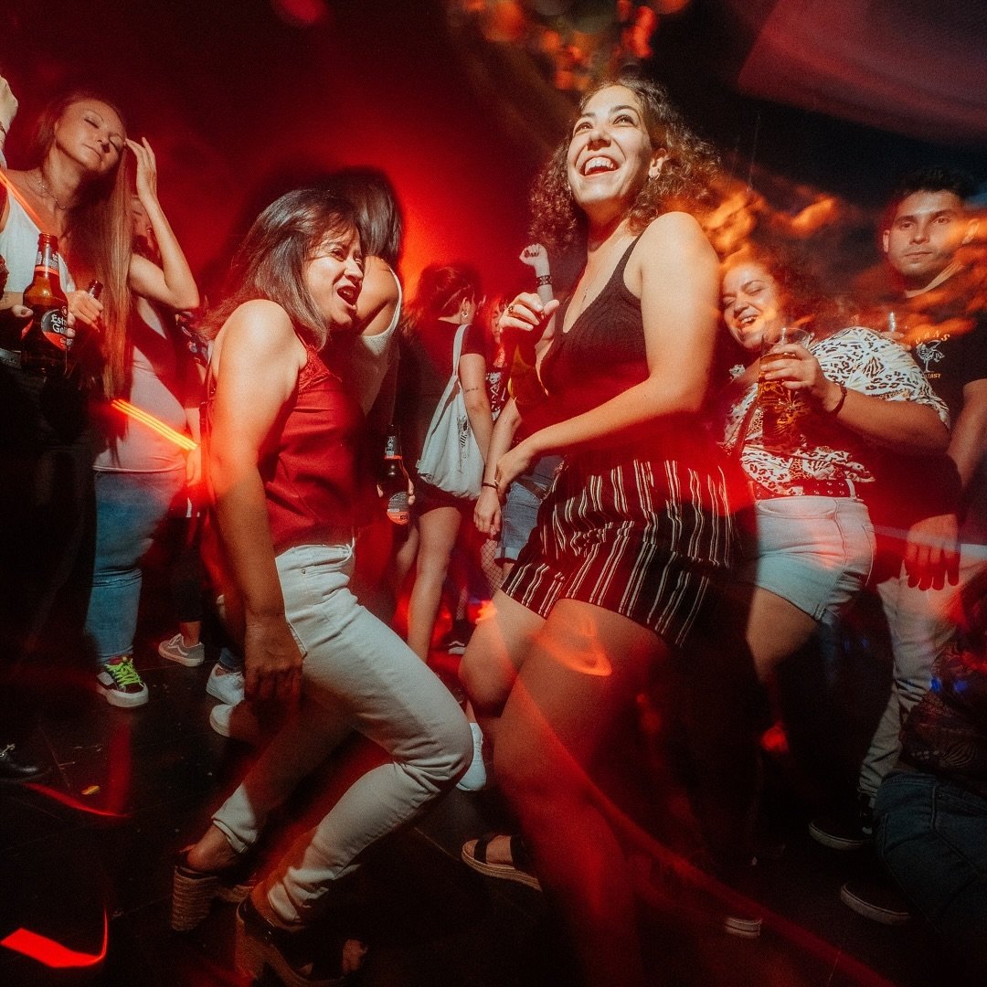 Grupo de personas bailando en una pista de baile con luces rojas y efectos borrosos. Dos mujeres en el centro, una de ellas con un top rojo y pantalones blancos, la otra con un vestido de rayas, se ríen y disfrutan la música.