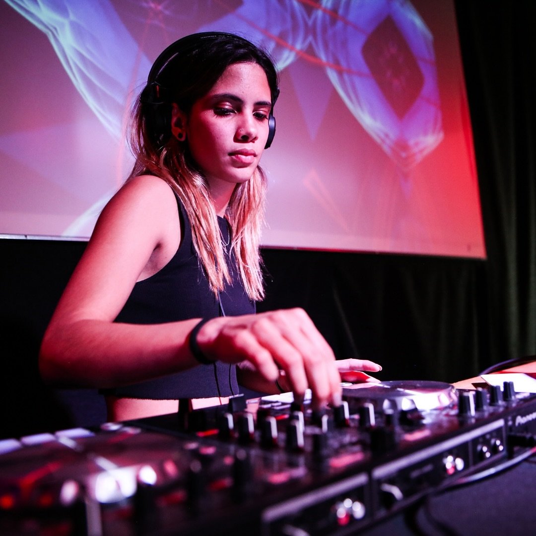 DJ mujer, concentrada, mezcla música en una consola con auriculares puestos. Detrás de ella, una pantalla proyecta gráficos de luces en tonos morados y rojos.