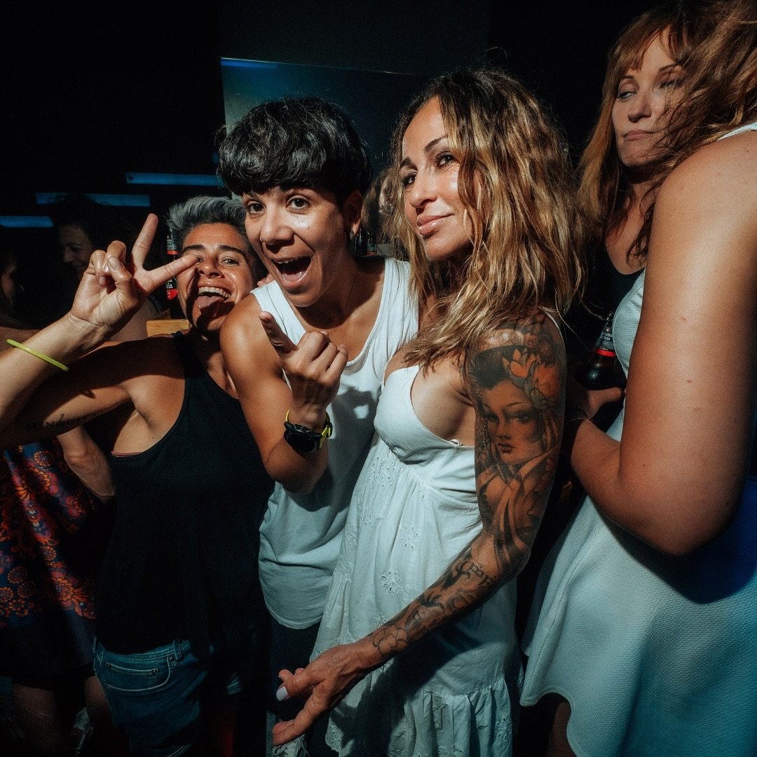 Tres personas posando en lo que parece ser una fiesta. Una mujer a la derecha con tatuajes visibles en su brazo, otra persona al centro haciendo un gesto de paz con la mano, y una más sonriendo ampliamente.