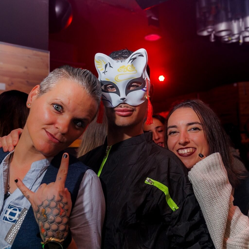 Tres personas posan juntas, una con el cabello corto y tatuajes en la mano haciendo el gesto de "cuernos". La persona en el centro lleva una máscara de gato decorativa. El fondo tiene luces rojas y se percibe una atmósfera festiva.