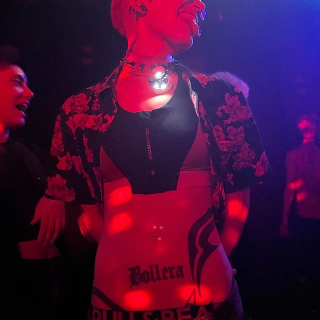 Persona con cabello corto teñido, luciendo un atuendo de estilo alternativo con tatuajes visibles en el abdomen que dicen "Bollera". Está en un ambiente oscuro con luces rojas que iluminan la escena.