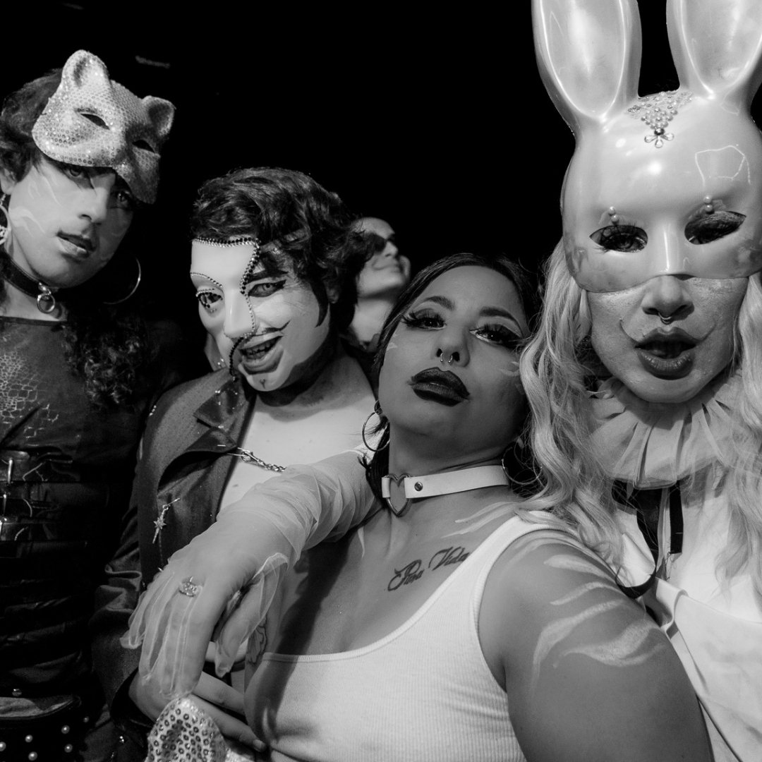 Cuatro personas enmascaradas posan para la cámara en blanco y negro. Las máscaras tienen diseños extravagantes, incluyendo una máscara de conejo y otra de gato.